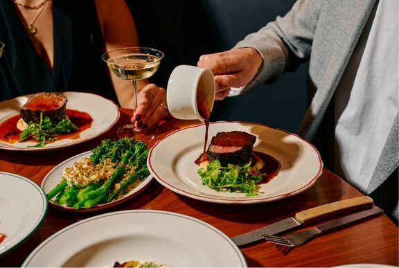 Chef pouring gravy onto a steak dish at a restaurant table.