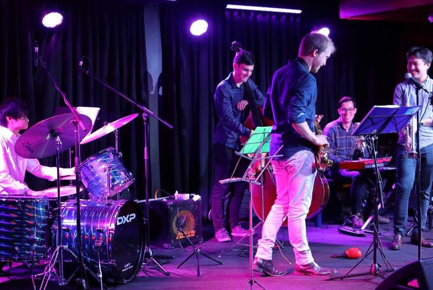 A group of musicians on stage performing under a pink light. 