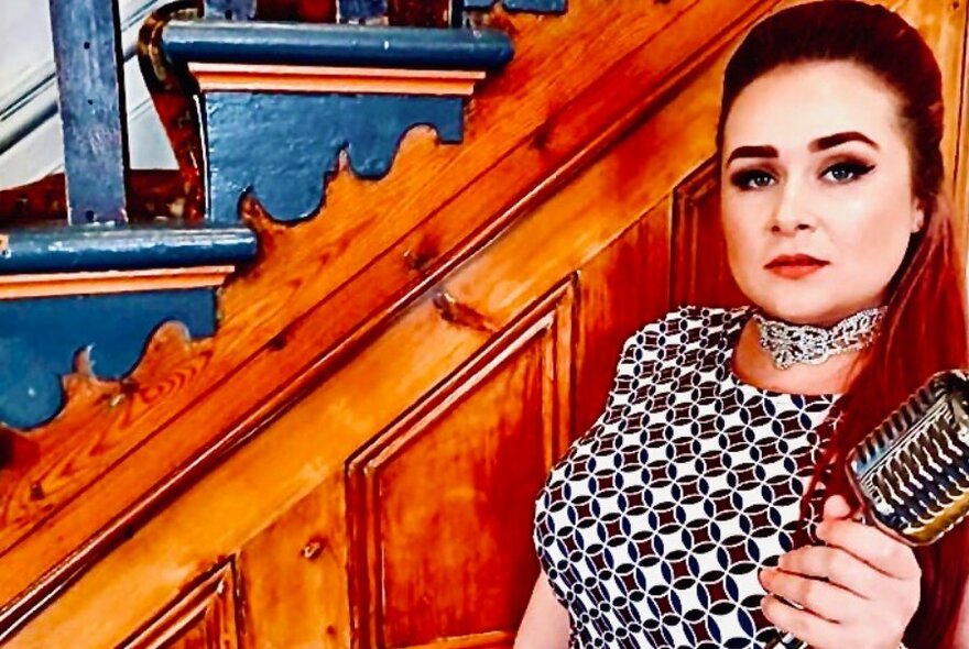 A woman in a 1960s-style dress holds a retro microphone near an ornate wooden staircase.