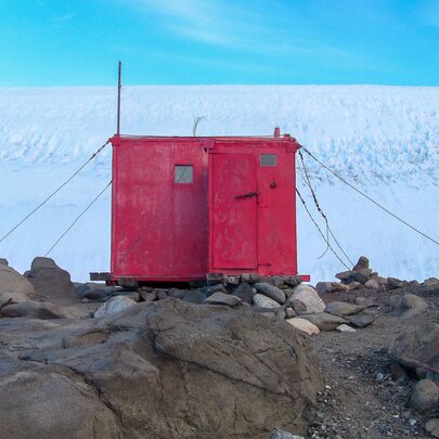 The red Platcha Hut at the edge of the Antarctic Plateau, from 2010, set on a rocky landscape with large areas of ice and blue sky in the background.