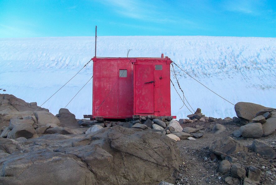 The red Platcha Hut at the edge of the Antarctic Plateau, from 2010, set on a rocky landscape with large areas of ice and blue sky in the background.