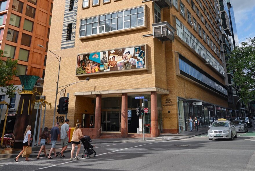The Hero Apartment building at the intersection of two Melbourne city streets, with an artwork on the cream brick facade above the entrance, with people crossing the road, a car and traffic lights.