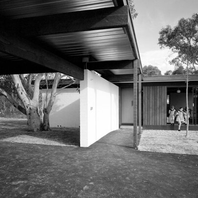 A mid-century modern house featuring a flat-roofed carport, a covered walkway with a privacy wall, and glass-panelled entry doors where people are sitting.