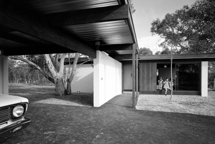 A mid-century modern house featuring a flat-roofed carport, a covered walkway with a privacy wall, and glass-panelled entry doors where people are sitting.