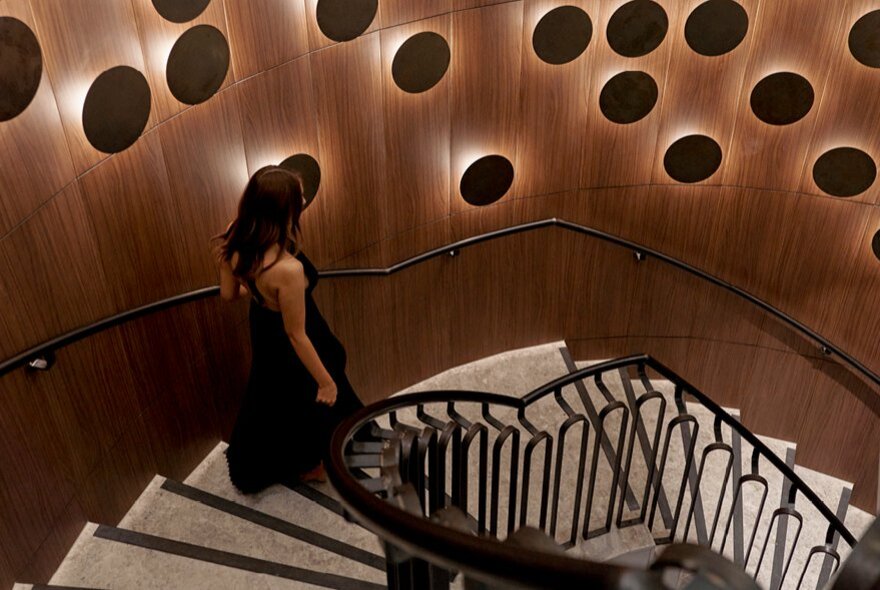 A woman in black descending a curved marble staircase, with circular black light fittings on the wooden wall beside it.
