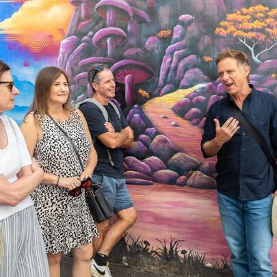 Small tour group listening to a man talking, holding a hat and standing in front of a wall mural.