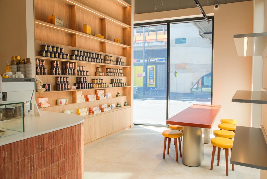 Shelving stocked with goods in a small cafe with a long bench and small yellow stools.