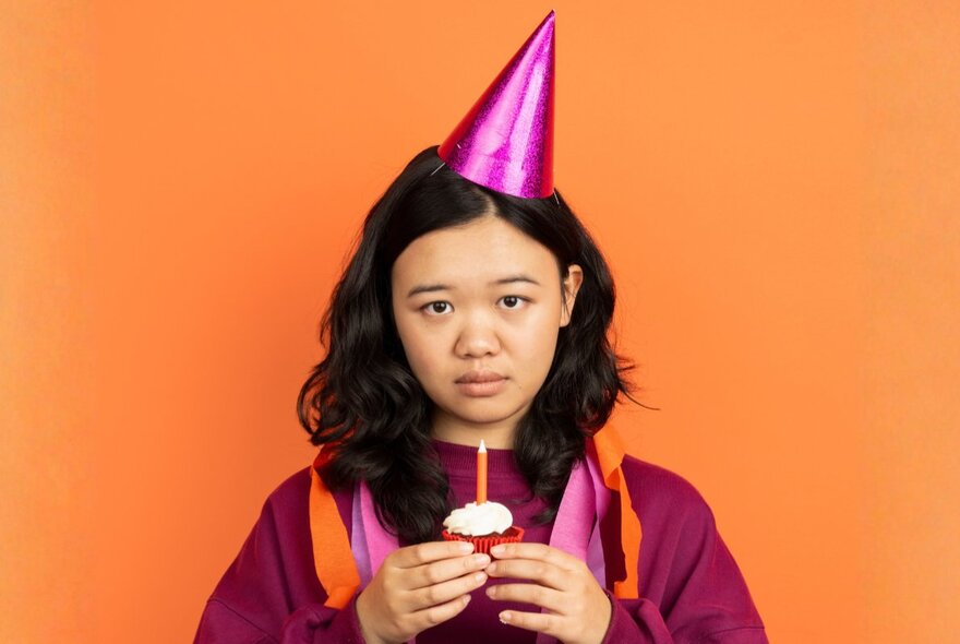 A young woman in a graduation robe holding a cupcake with a single candle, wearing a pink party hat and a sad expression.