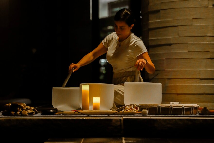 A person plays crystal singing bowls on a low table illuminated by warm candles as part of a sound bath immersive experience.