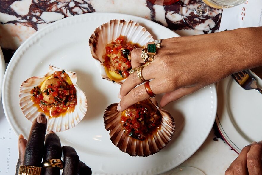 Two people's hands each taking a stuffed scallop off a white plate.