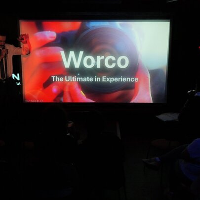 A screen on a small stage showing a power-point display with the text 'Worco: The Ultimate in Experience' with a blurry visual of someone holding a camera in the background, and a presenter speaking into a microphone standing on the stage in a darkened venue.