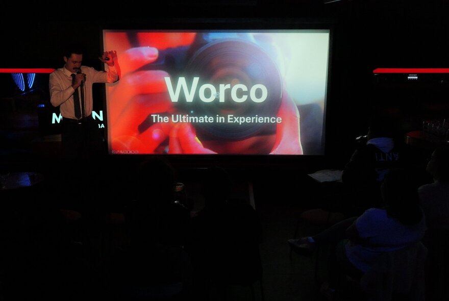 A screen on a small stage showing a power-point display with the text 'Worco: The Ultimate in Experience' with a blurry visual of someone holding a camera in the background, and a presenter speaking into a microphone standing on the stage in a darkened venue.