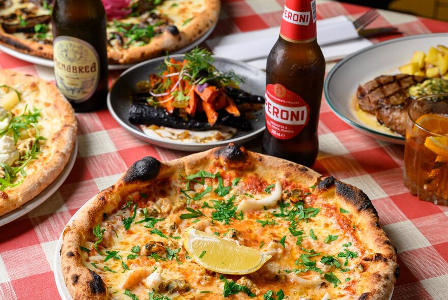 Plates of pizza with a salad and two beers on a red and white checked tablecloth.