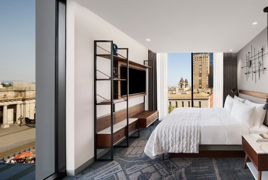 A hotel room with a white linen covered bed, patterned carpet and large windows.