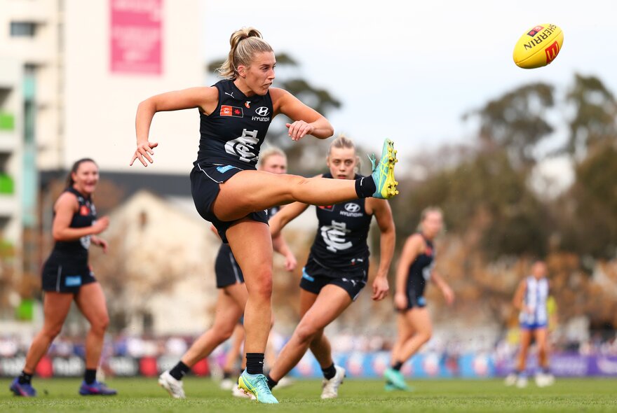 A Carlton AFLW player mid-kick with her leg raised high and the yellow football travelling through the air, other players in the background.