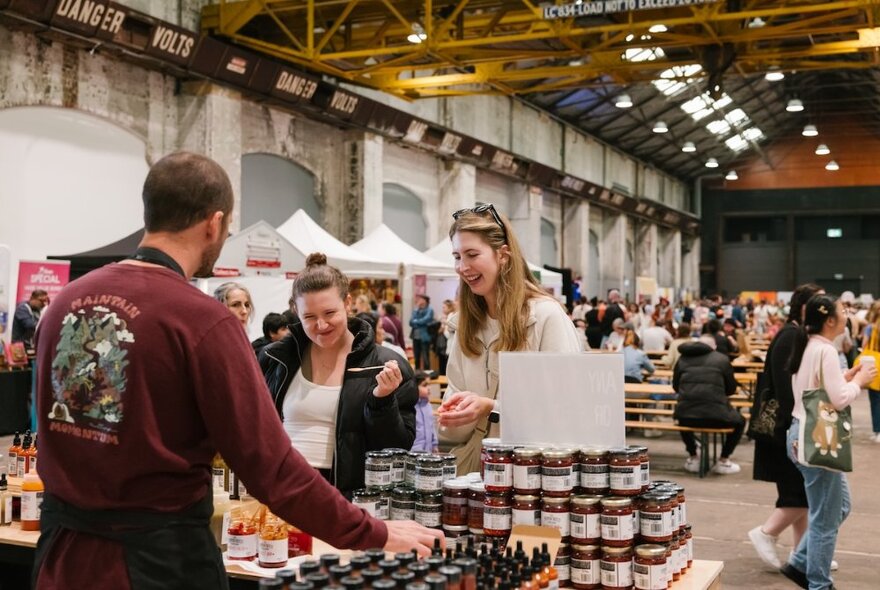 A market stall holder with food products, engaging with two people at the stall who are sampling the products, people walking around other stalls in the background.