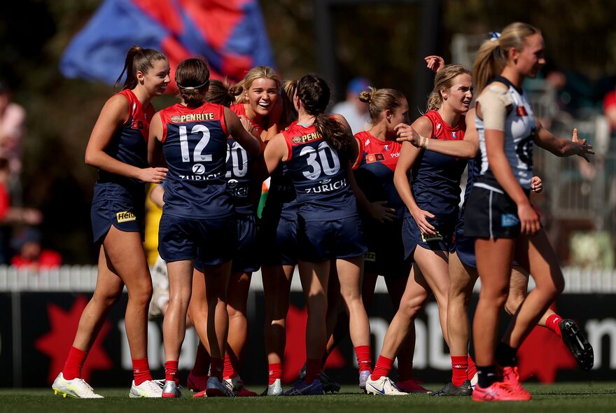Melbourne AFLW players celebrating on the field, coming together for a group huddle.