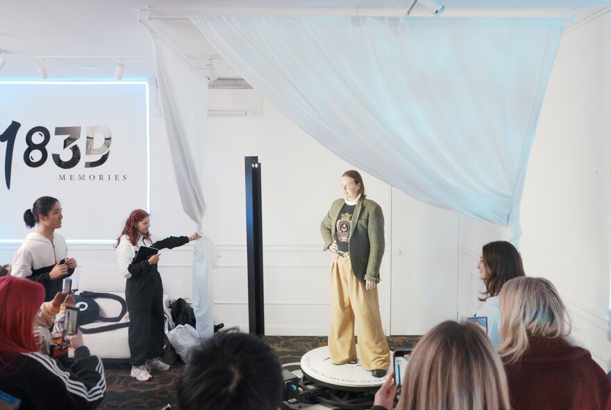 A group of people standing and watching as a woman on a small pedestal is photographed near a white curtain. 