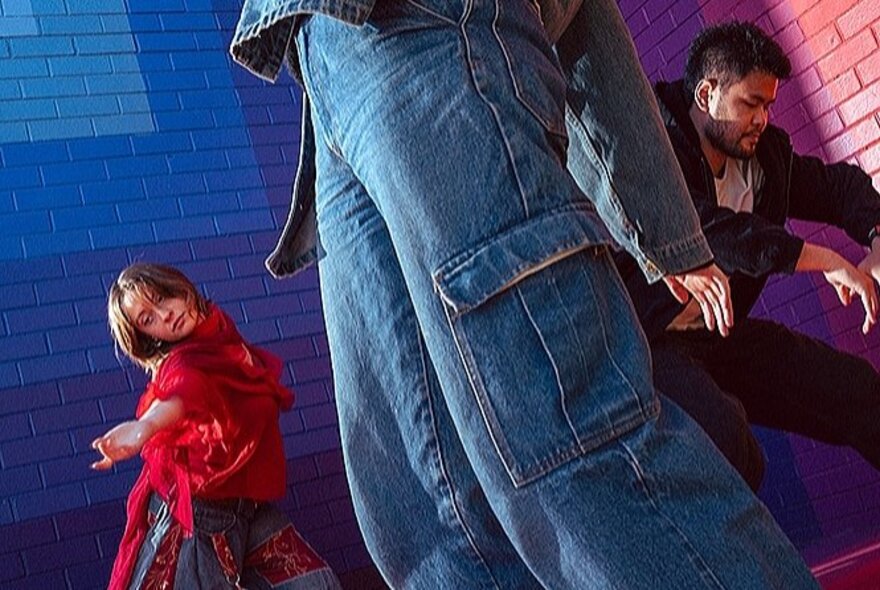 The lower half of a person's body, dressed in cargo jeans, while a woman and a man dance separately in the background in front of a rainbow-coloured brick wall.