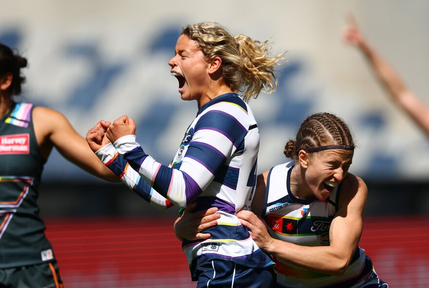 A Geelong Cats AFLW player celebrating with a double fist pump during a game while a teammate hugs her by the waist.