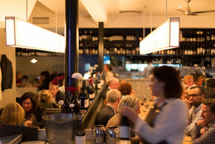 View across busy restaurant, with wine shelving at rear, and waiter in foreground.