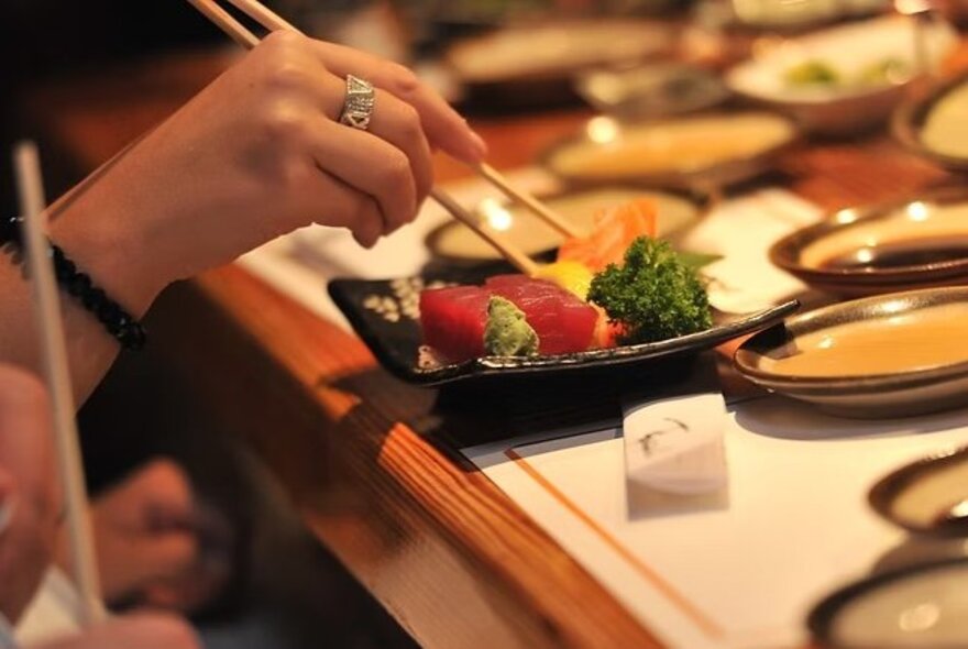 A woman's hand holding chopsticks as she picks up a slice of sashimi from a traditional Japanese plate surrounded by other small Japanese crockery.