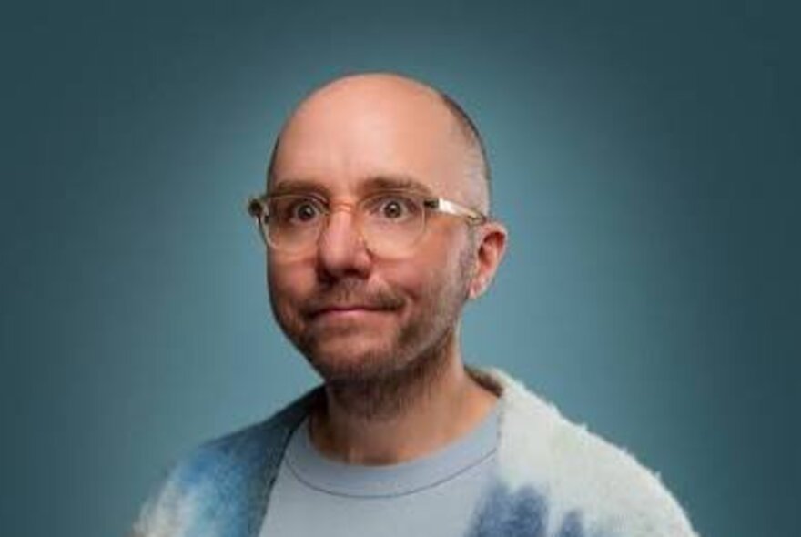 Comedian Tommy Dassalo, a middle-aged bald man wearing glasses and a blue top, he has a bemused expression and is posed against a teal background.