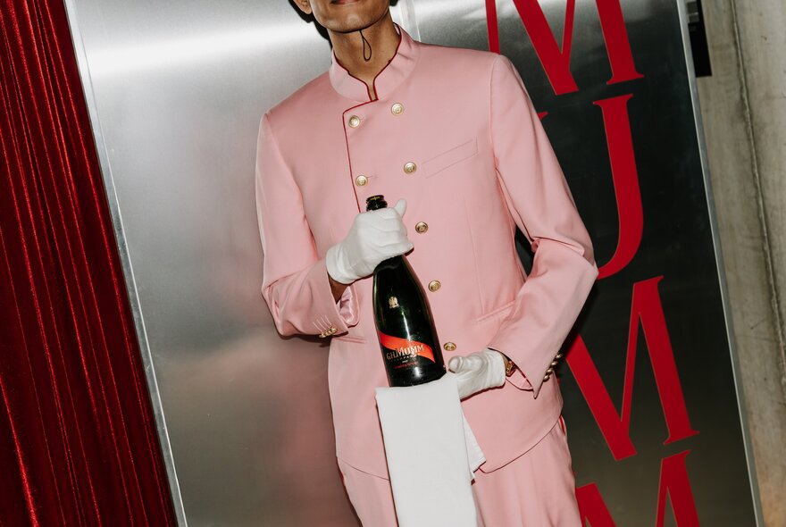 A waiter in a pink suit holding a bottle of Mumm champagne and a white serving napkin.