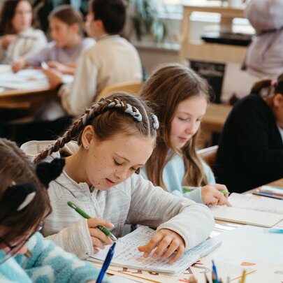 Kids seated around large tables writing into notebooks during a creative writing workshop in a bright studio space