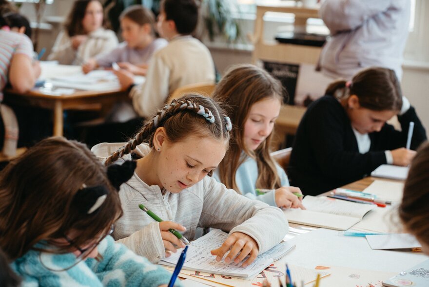 Kids seated around large tables writing into notebooks during a creative writing workshop in a bright studio space