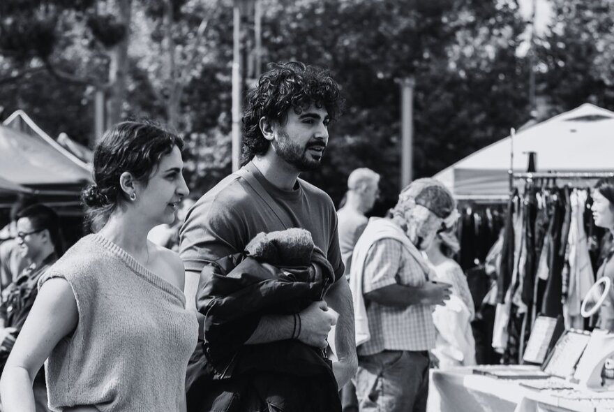 A black and white image of two people looking at a stall at an outdoor market. 