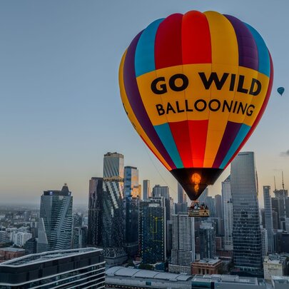 A hot air balloon in rainbow stripes with GO WILD Ballooning written on it floating above Melbourne, another balloon in the background. 