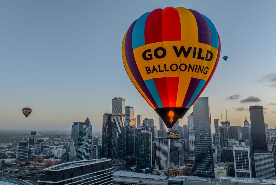 A hot air balloon in rainbow stripes with GO WILD Ballooning written on it floating above Melbourne, another balloon in the background. 