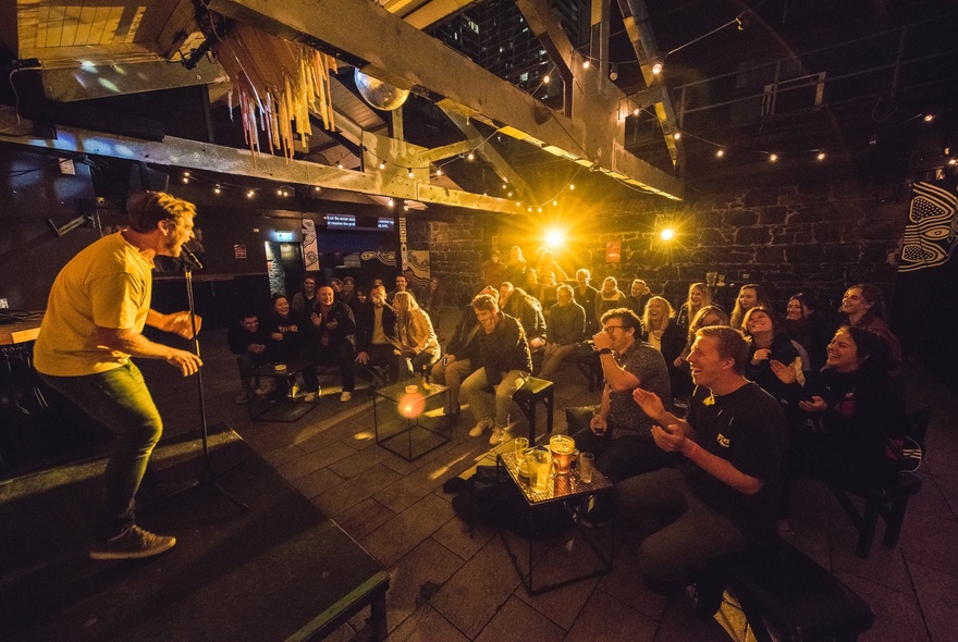 Comedy performer holding a microphone on stage with cheering audience looking on.