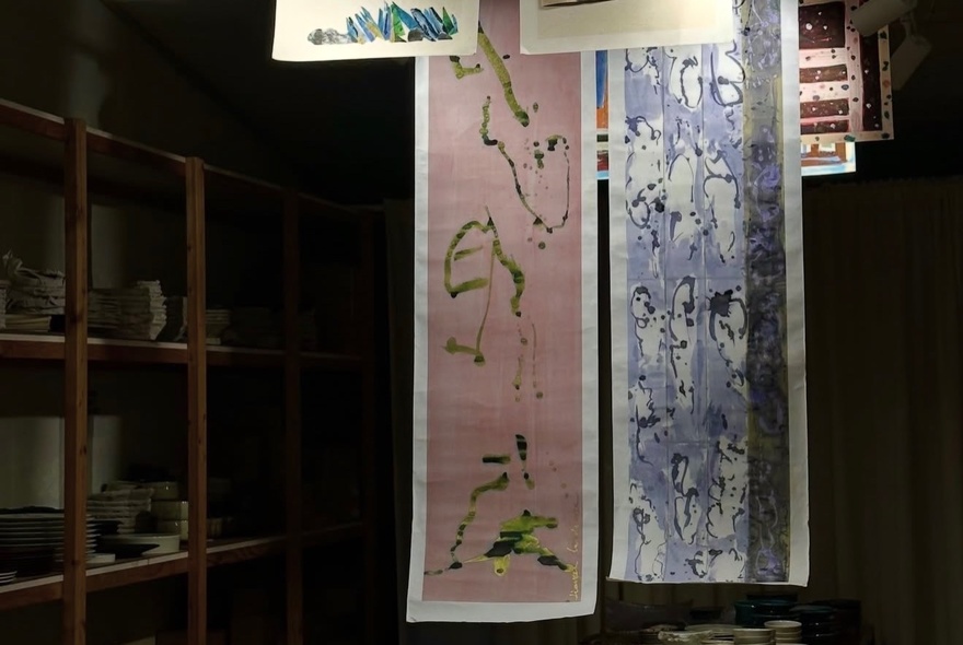 Two long hanging scrolls (kakemono) displayed in a dimly lit gallery studio next to a wooden shelving unit.