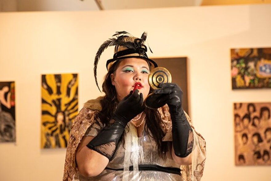 A person holding up a small compact mirror and applying lipstick to her mouth, wearing black elbow length gloves and a hat with feathers, behind her are paintings on  a gallery wall.