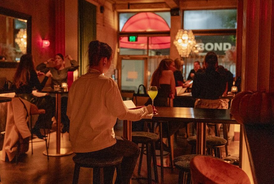 Interior of a bar with soft lighting and people seated at high wooden tables and chairs.