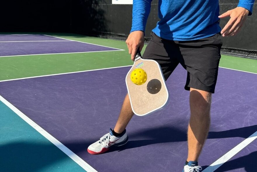 A person on a pickleball court, just the lower half of their body visible, holding a wooden paddle used to play pickleball and hitting a ball with it.