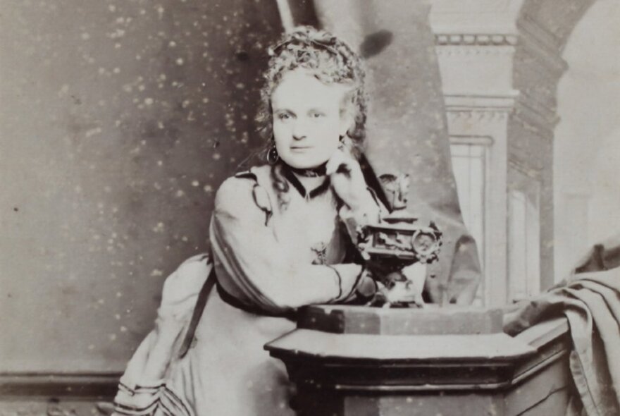 Victorian-era figure Madame Brussels, leaning on a photographer's pillar with her hand to her chin, wearing an elaborate gown.