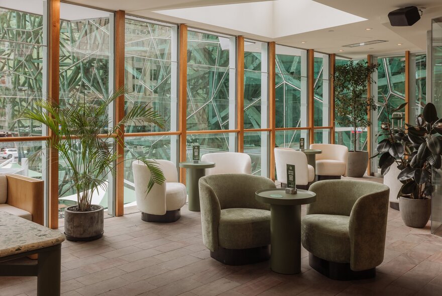 The lounge area at Good Nature Hotel with club armchairs and small round tables, potted plants and large floor to ceiling windows.