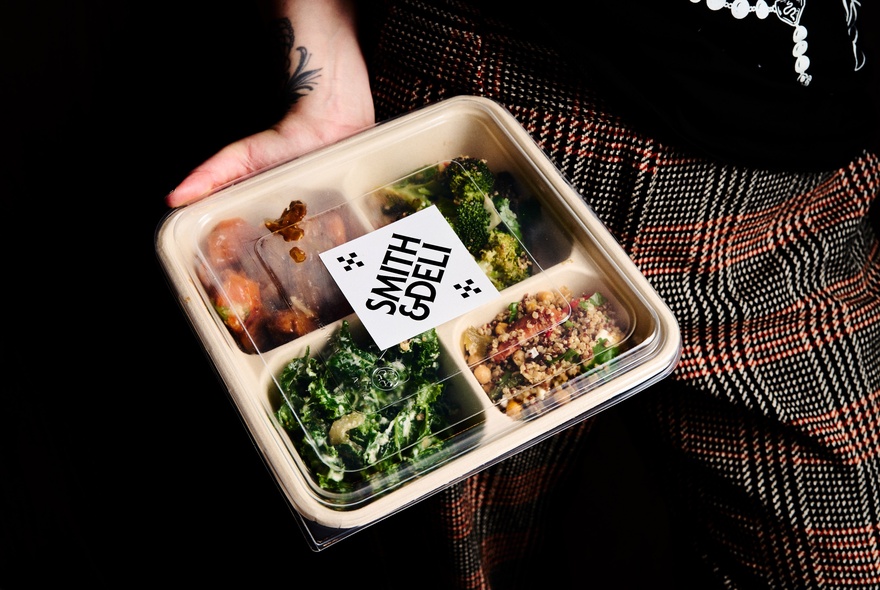 Hand holding a Smith + Deli takeaway bento-box style container with four portions of food.