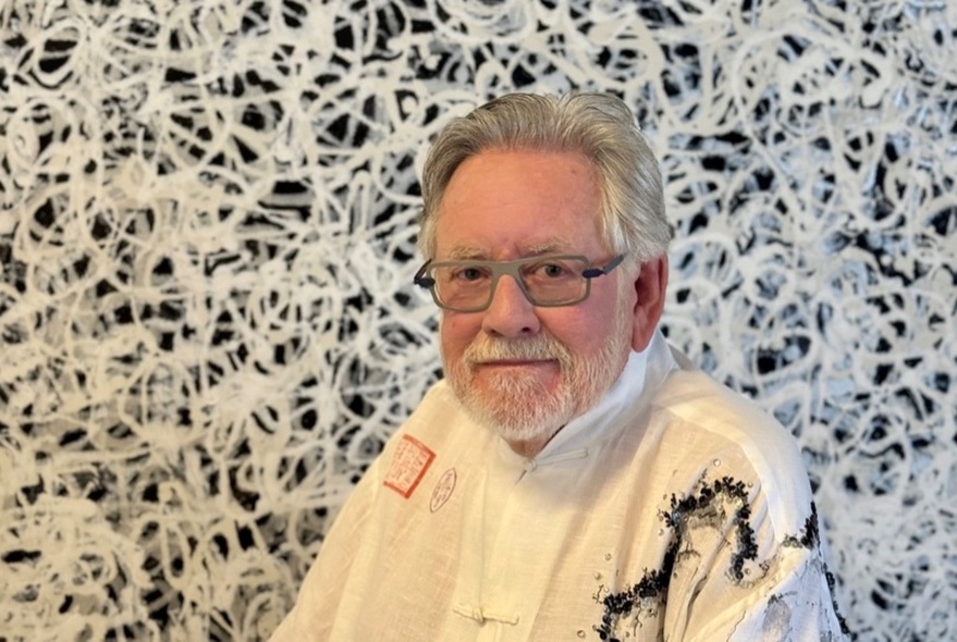 Artist Gregory Ladner, a middle-aged man wearing a white shirt and glasses, posed in front of one of his abstract paintings.