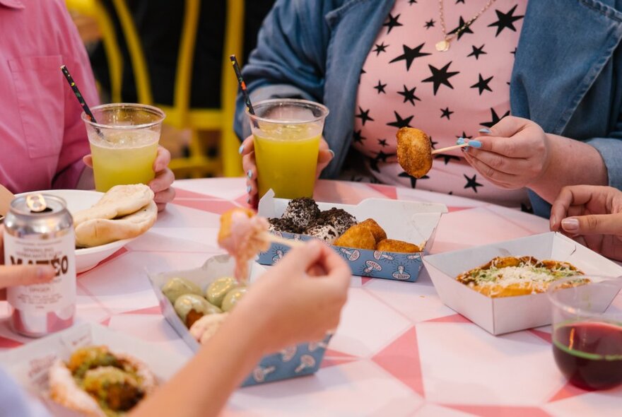 Hands selecting food with small forks from takeaway food containers arranged on a round  table.