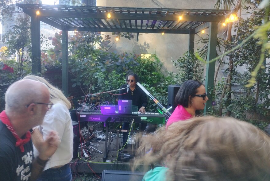 Soul musician Dinash Daniel playing the keyboard in an outdoor setting with people dancing and moving in the foreground. 