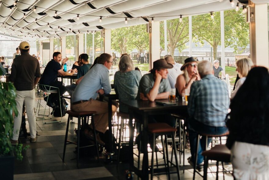 People seated on stools at tall bar tables on an outdoor restaurant terrace with leafy trees in the background. 