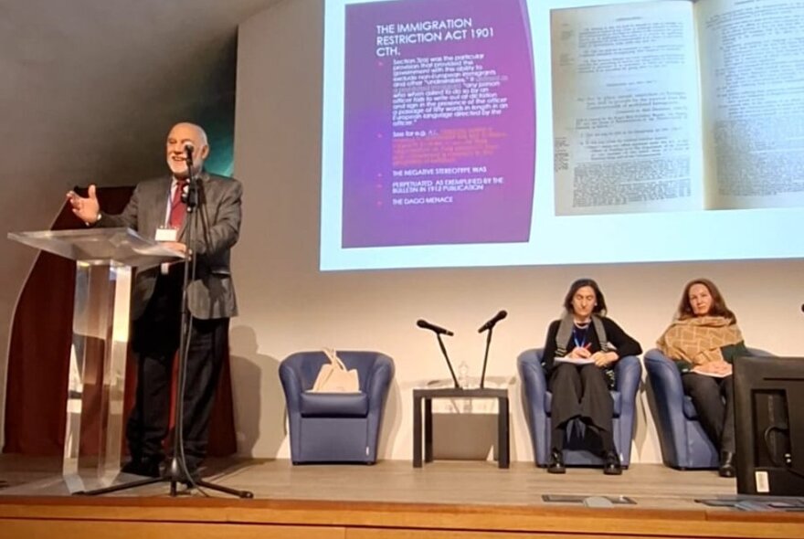 A speaker at a podium on stage, with a projected image on the wall and two women seated in chairs behind him.