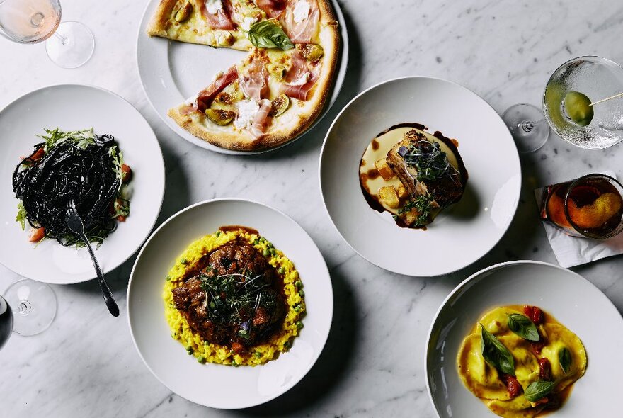 Overhead view of several dishes on white plates on a marble surface including pizza and pasta.