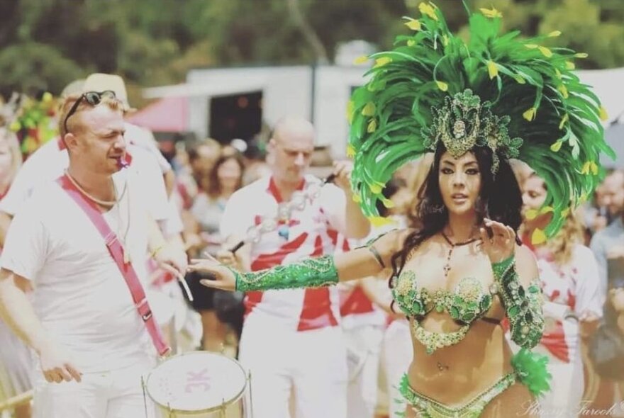 A woman wearing a Latin American dancing costume with a large green feather headdress, dances in front of a line of men in white uniforms, one wearing a drum.