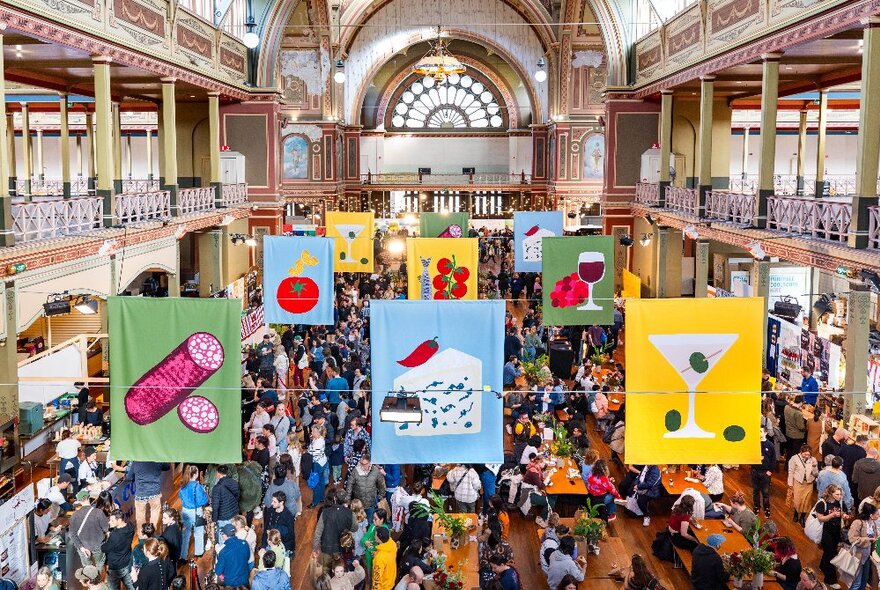 Aerial view of large crowds of people walking around The Little Food Market at the Royal Exhibition Building, with colourful banners hanging from the ceiling.