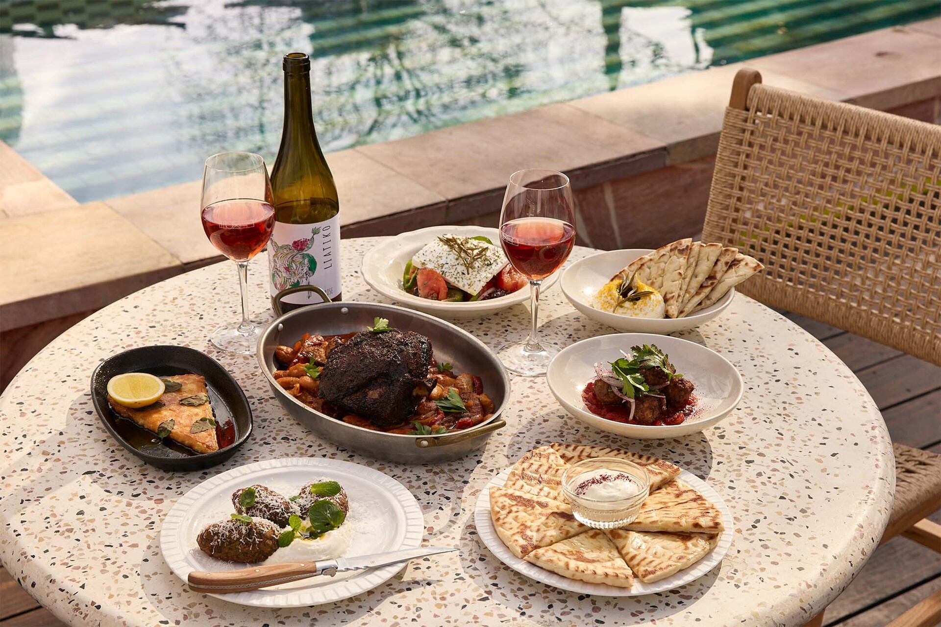 A table at an outdoor restaurant next to a pool with plates of Greek food and wine on it.